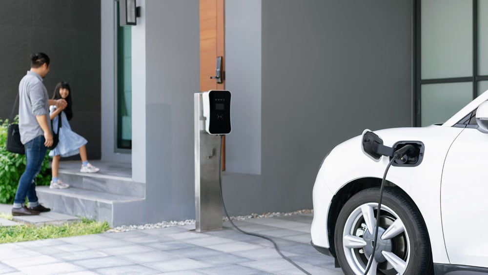 Ein weißes Elektroauto lädt vor einem Wohnhaus an einer Wallbox, im Hintergrund ein Mann mit Kind – symbolisiert datenbasierte E-Auto-Beratung und die Berücksichtigung individueller Mitarbeiterbedürfnisse