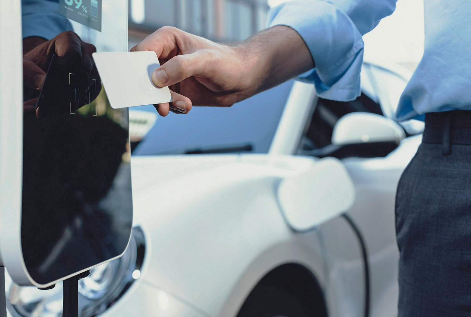 Mann hält eine RFID-Karte vor eine moderne Ladesäule, während im Hintergrund ein weißes E-Auto geparkt ist – Darstellung für flexible und kompatible Ladehardware-Lösungen im Unternehmenskontext