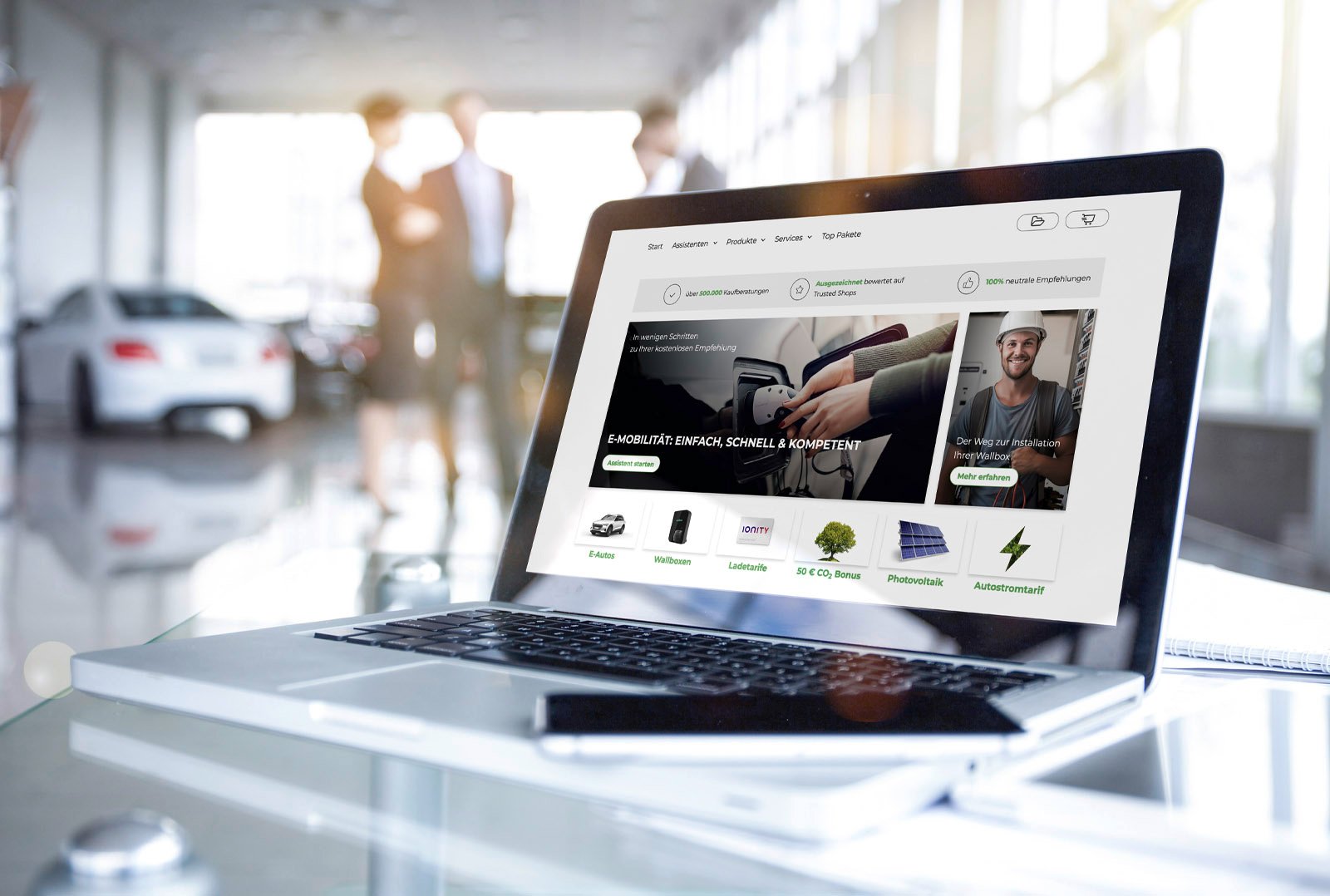 Laptop mit geöffneter e-Kaufberater-Oberfläche auf einem Glastisch im Autohaus, im Hintergrund mehrere Personen im Gespräch