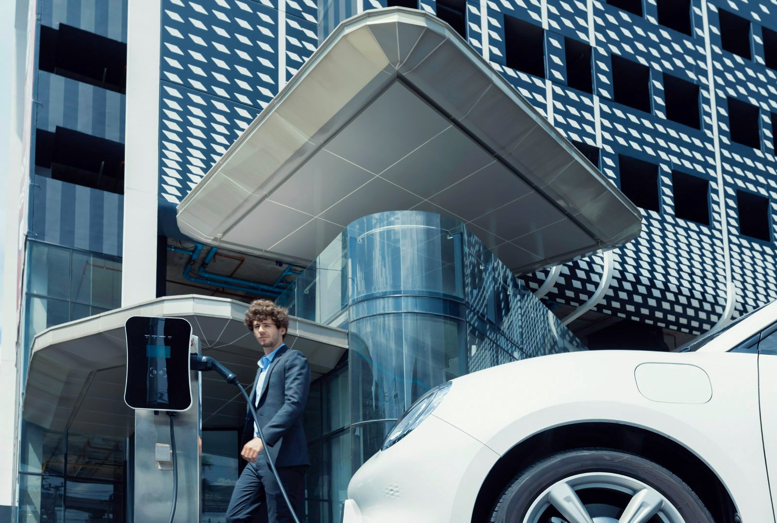 Mann im Anzug steht neben einer Ladesäule vor einem modernen Autohaus und lädt ein Elektroauto – Symbol für Standortelektrifizierung und moderne Ladeinfrastruktur im Autohaus